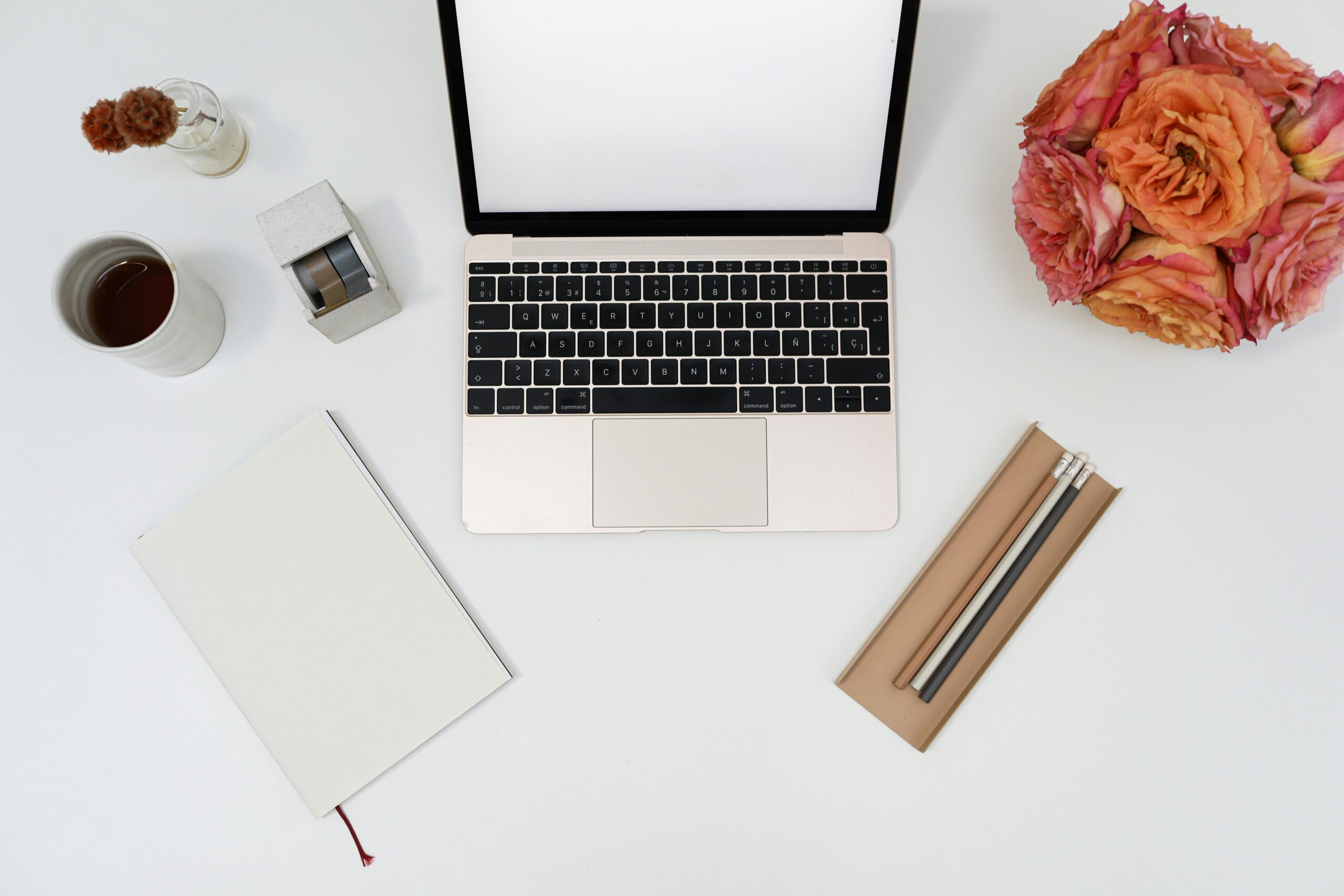A top view of a minimalistic workspace featuring a laptop, flowers, and stationery on a clean desk.